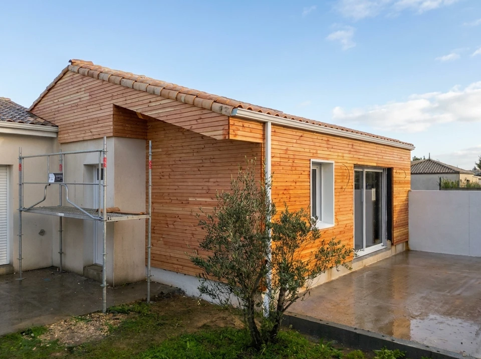 Vue d'une maison agrandie par une extension en ossature bois. La photo montre l'extension bois nouvellement construite accolée à la maison existante, avec les structures bois visibles ou partiellement couvertes, illustrant clairement l'ajout de surface habitable. L'extension doit être claire et bien intégrée à la construction existante, photographiée de jour en lumière naturelle, de façon à valoriser la transformation et l'agrandissement de l'espace de vie.