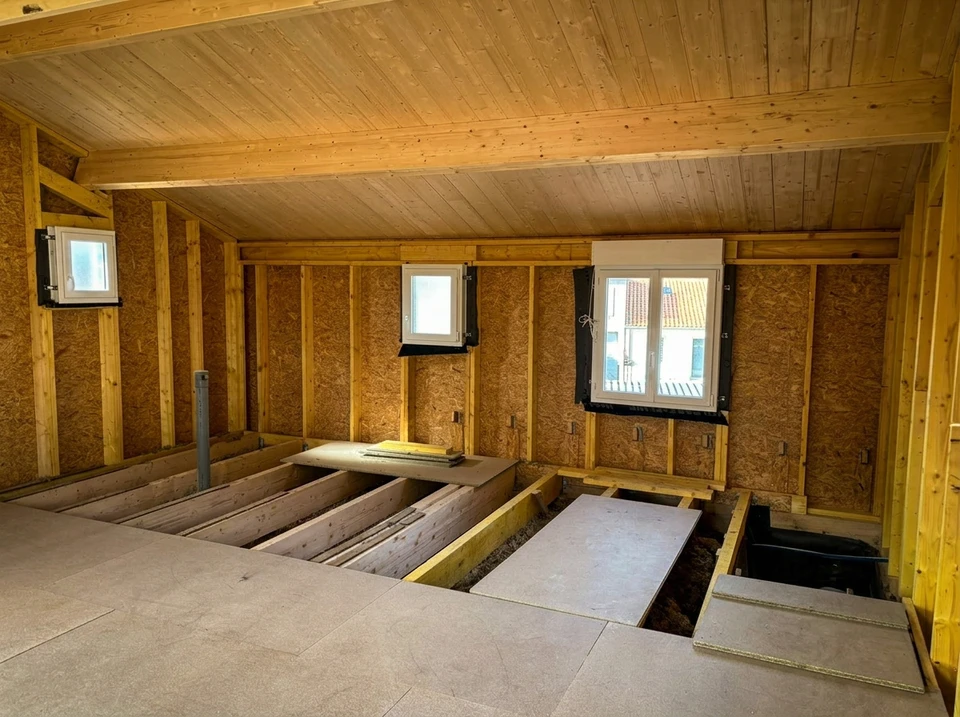 Ouvrier installant un système électrique dans une construction en bois, travaillant depuis un escabeau dans un espace intérieur avec ossature bois et isolation