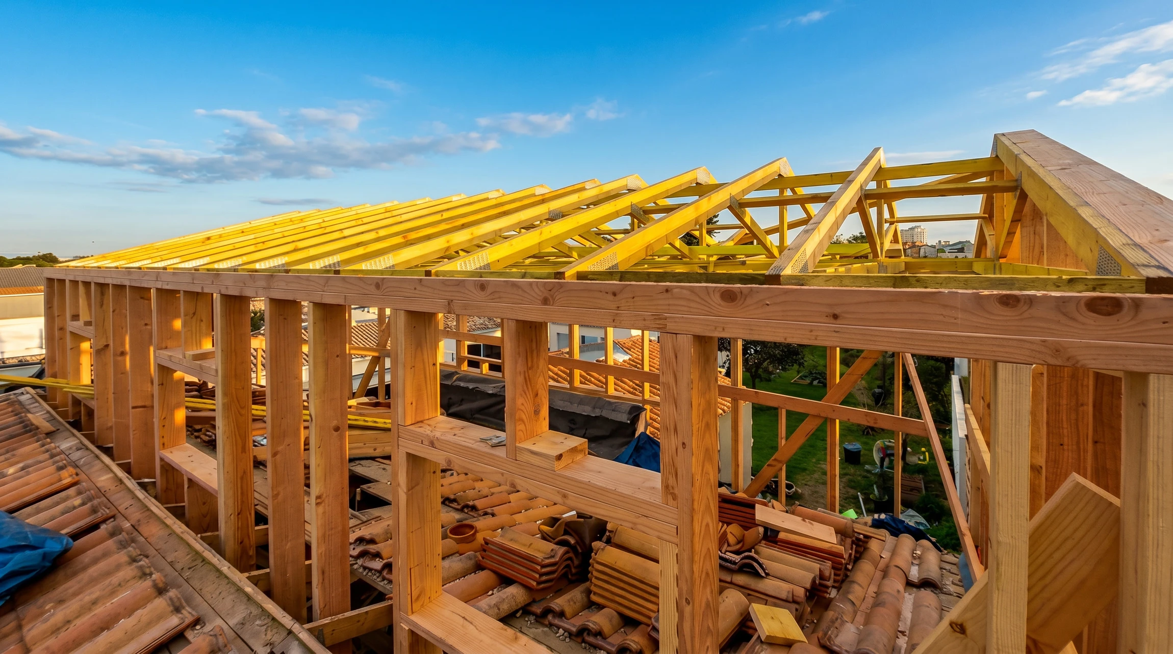 Chantier de construction résidentielle en cours, structure de charpente en bois apparent sur murs en briques rouges, ciel bleu avec nuages blancs en arrière-plan